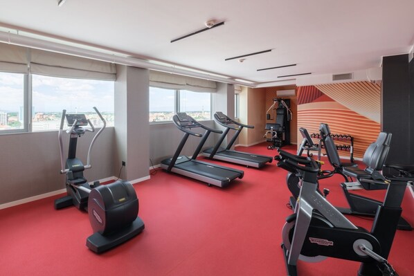 A gym with treadmills, an elliptical, and weight machines, large windows, and a red carpeted floor.