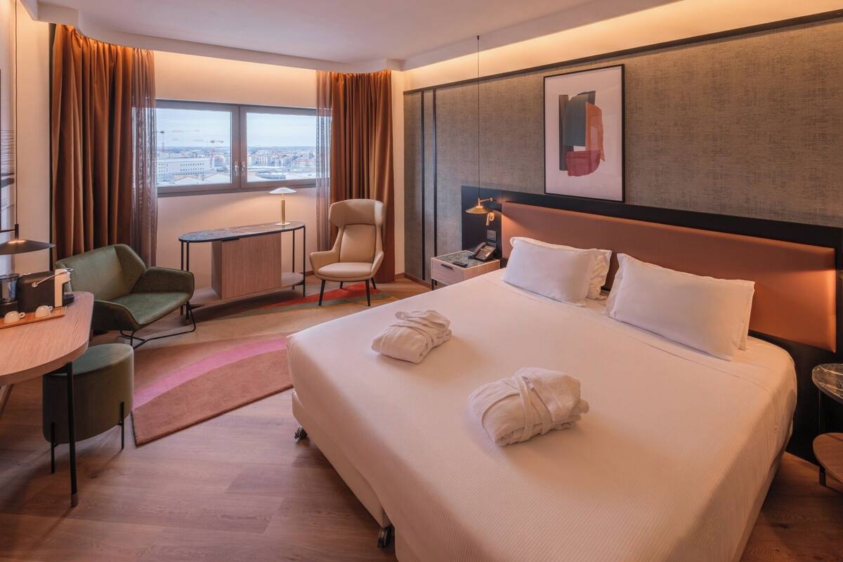 A hotel room with a large bed, a desk, a chair, a small table, and a window with curtains.