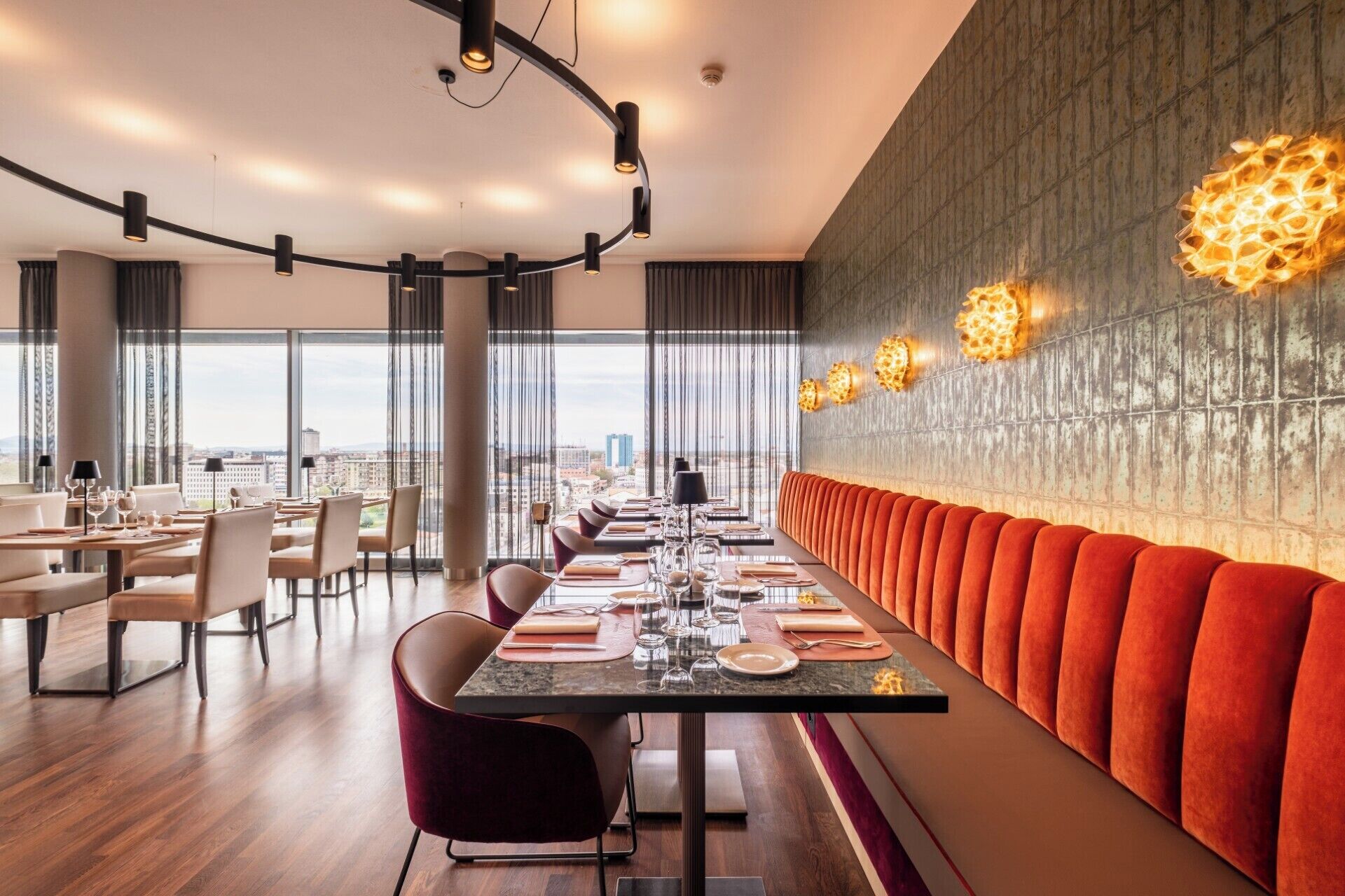 A modern dining area with a long table, cushioned seating, and a city view.