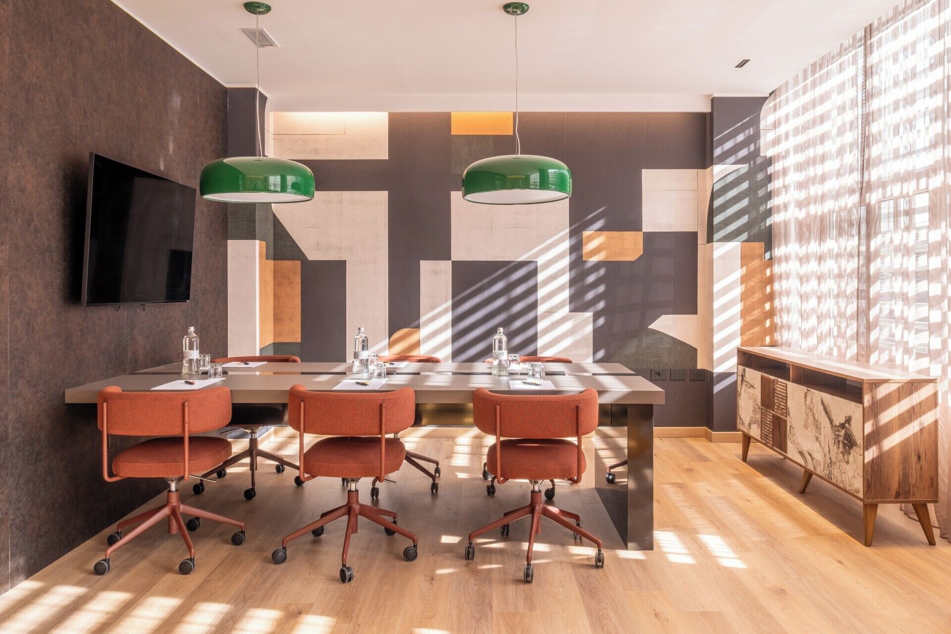 Uma sala de conferências com uma mesa comprida, quatro cadeiras laranja, uma TV de tela plana e arte geométrica nas paredes.