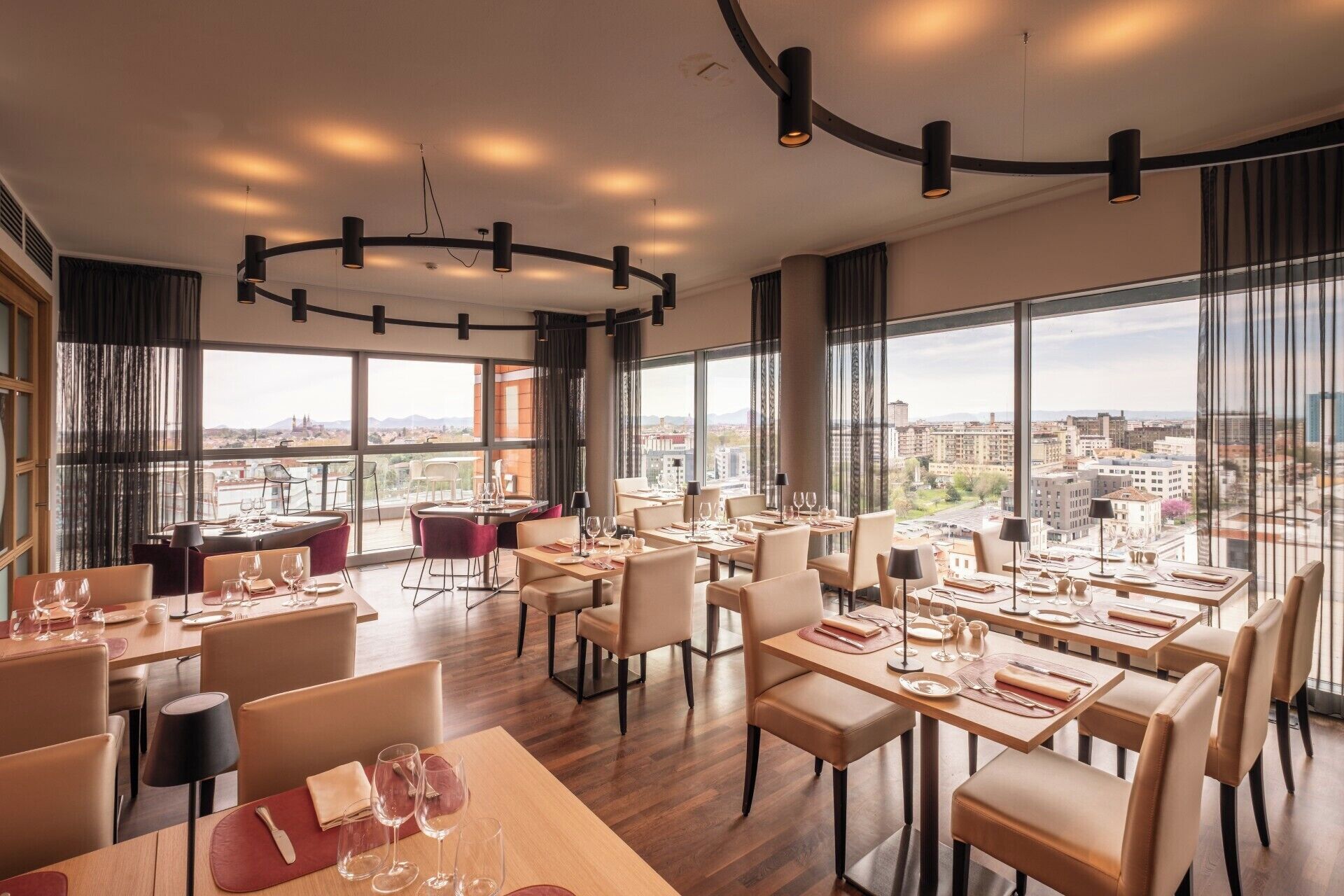 A modern dining area with tables set for dining, large windows offering city views, and a curved ceiling light fixture.