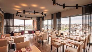 A modern dining area with tables set for dining, large windows offering city views, and a curved ceiling light fixture.