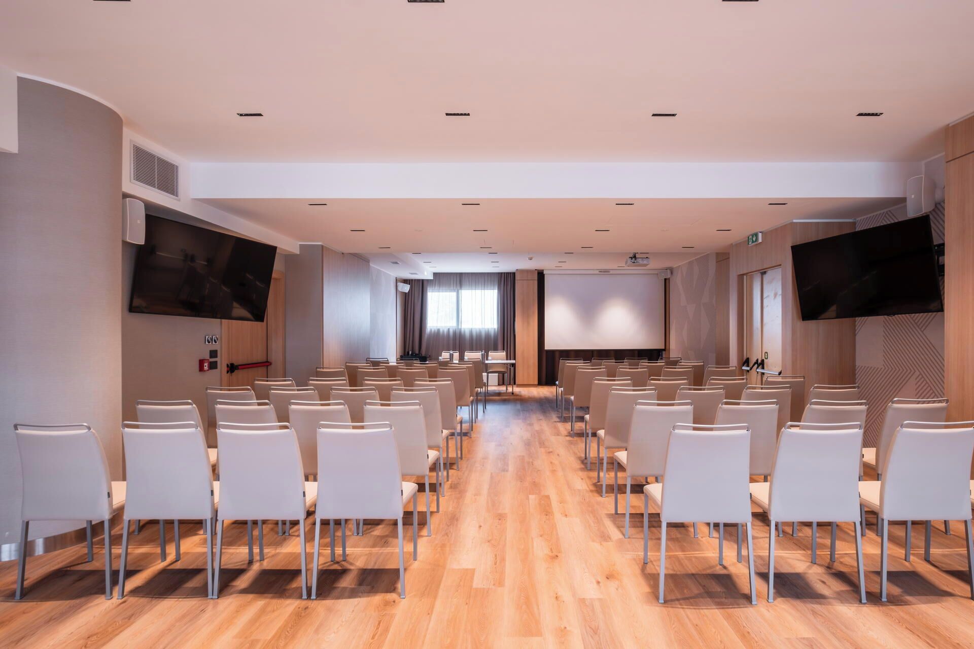 A conference room with rows of white chairs, a large screen at the front, and a wooden floor.