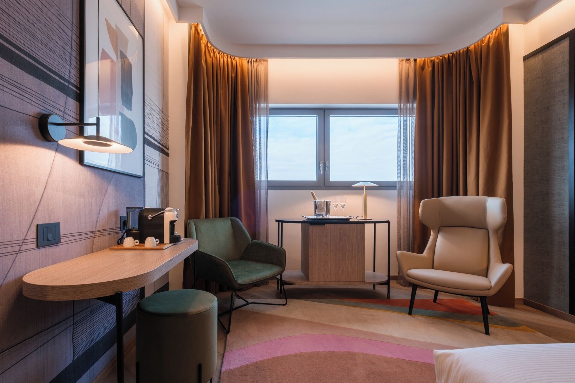 A hotel room with a desk, two chairs, a bed, a small table with a lamp, and a window with curtains.