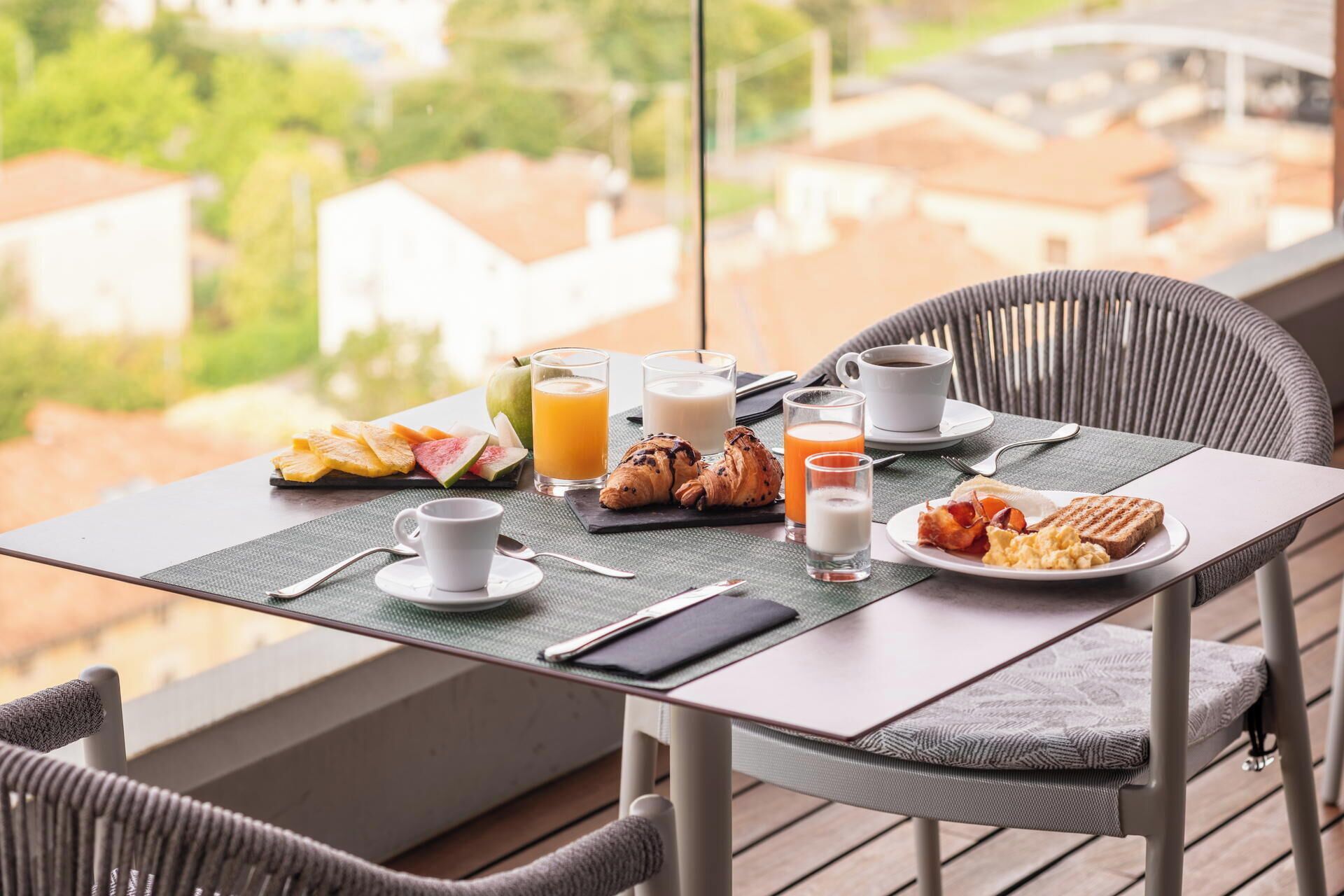 Una mesa de desayuno con cruasanes, fruta, jugo, café y un plato de huevos y tocino en un balcón con vistas a una zona residencial.