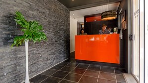 Una zona de recepció amb paret de pedra, una planta en test i un mostrador de color taronja vibrant.