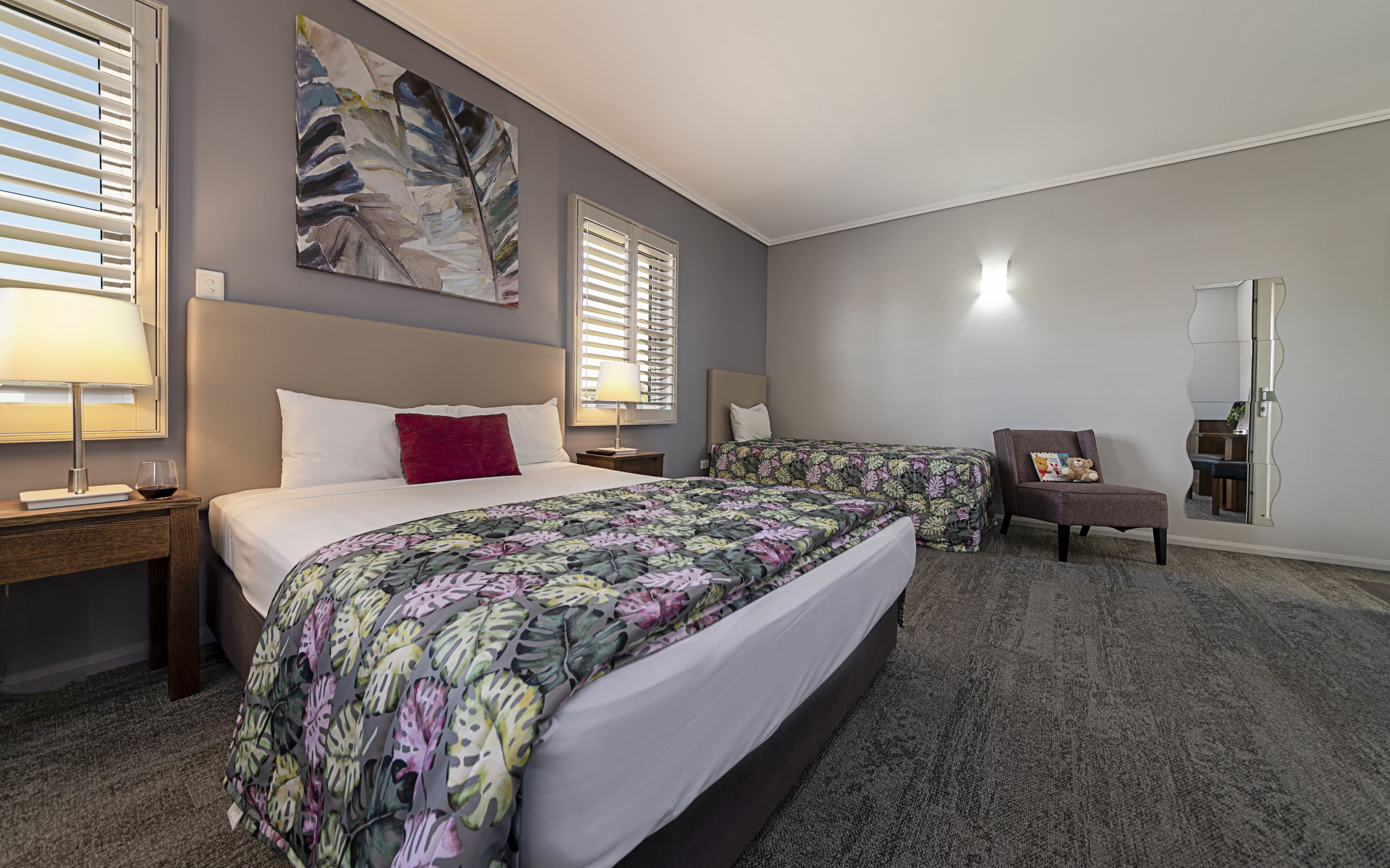 A hotel room with two beds, a large abstract painting, a mirror, and a chair with a teddy bear.