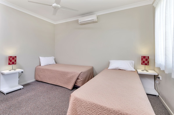 A hotel room with two single beds, a ceiling fan, an air conditioning unit, bedside lamps, and a carpeted floor. - Coachman's Inn Warwick (Warwick)