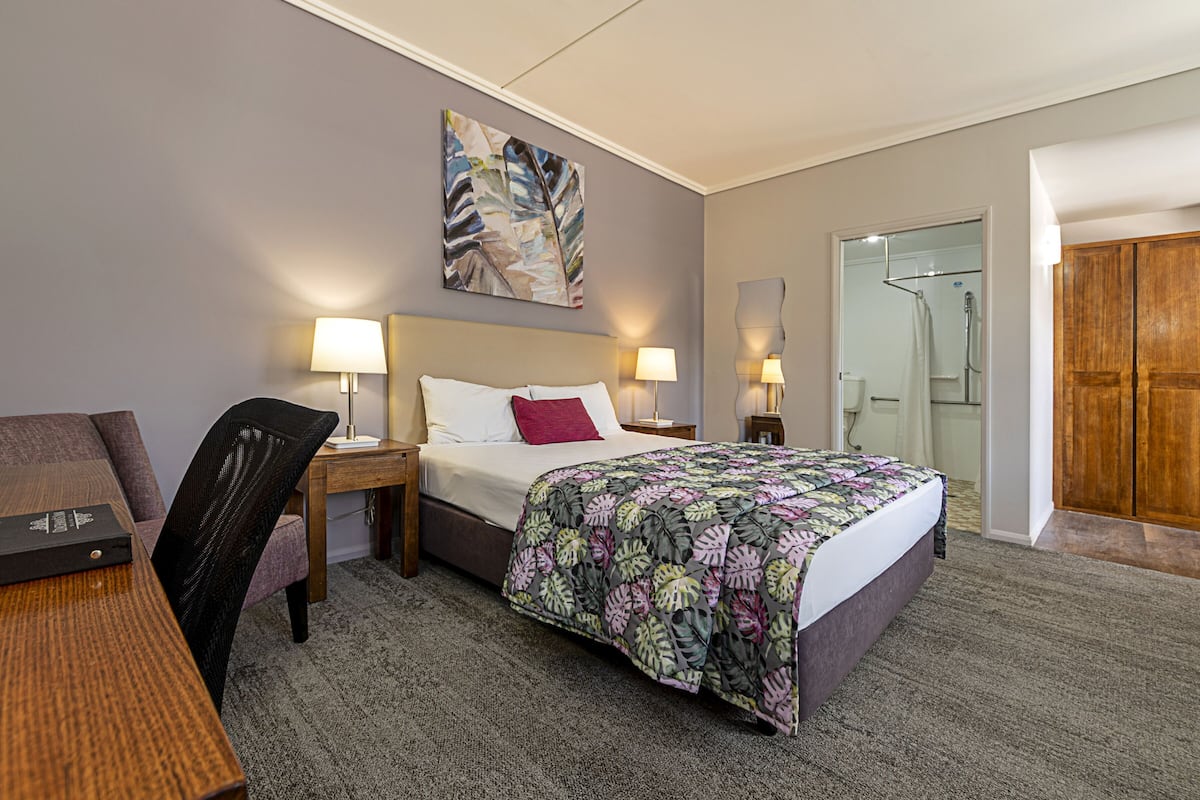 Habitación de hotel con cama, dos mesitas de noche con lámparas, una silla, escritorio, un cuadro en la pared y armario de madera.