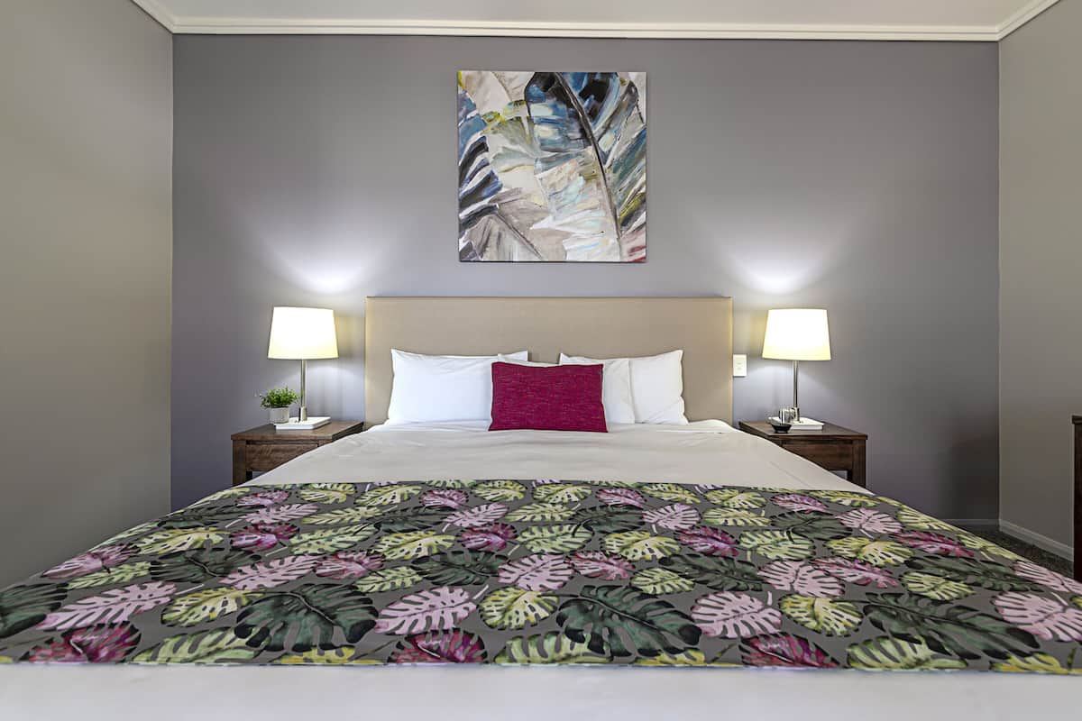 Una habitación de hotel con una cama, dos lámparas de noche, una mesita de noche, un cuadro abstracto en la pared y una colcha de diseño floral.