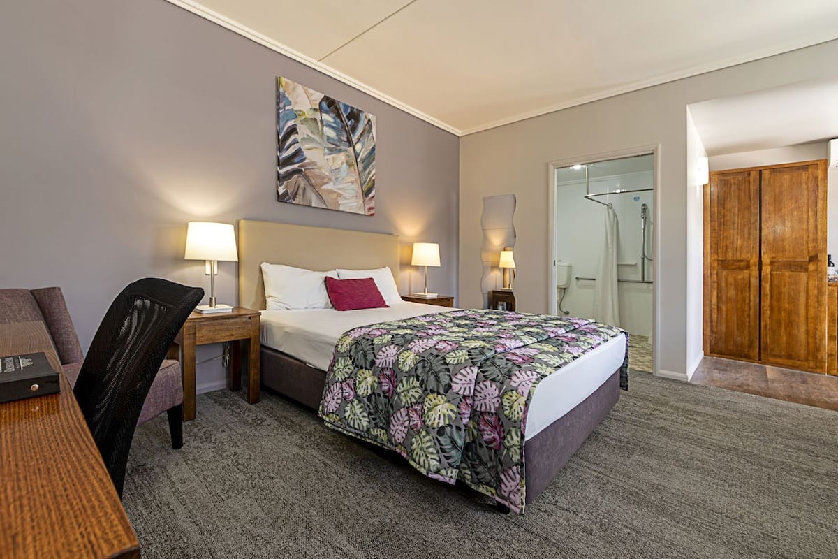 Habitación de hotel con cama, dos mesitas de noche con lámparas, una silla, un escritorio, un cuadro en la pared y un armario de madera.
