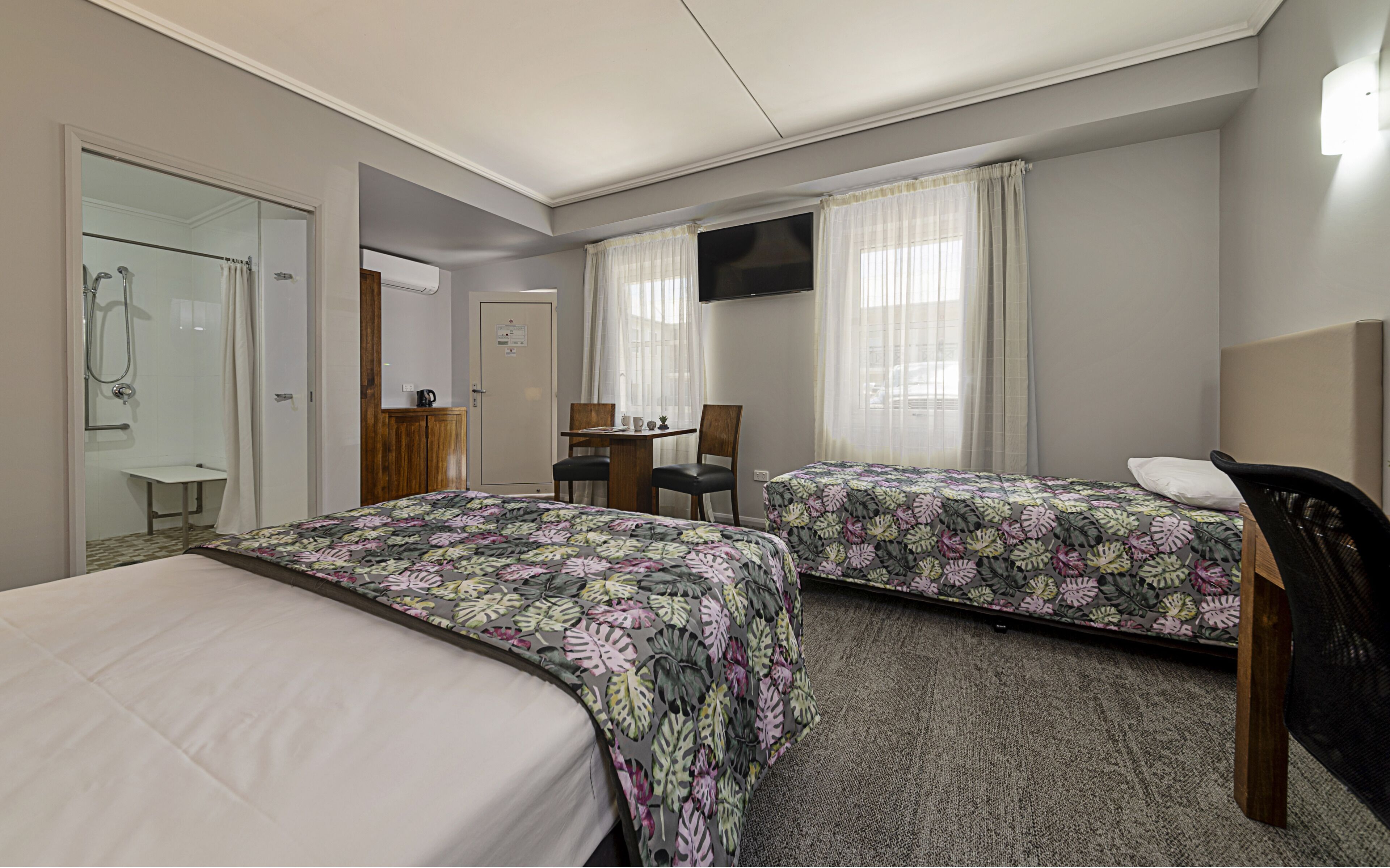 A hotel room with two beds, a desk, a chair, a TV, and a shower area.
