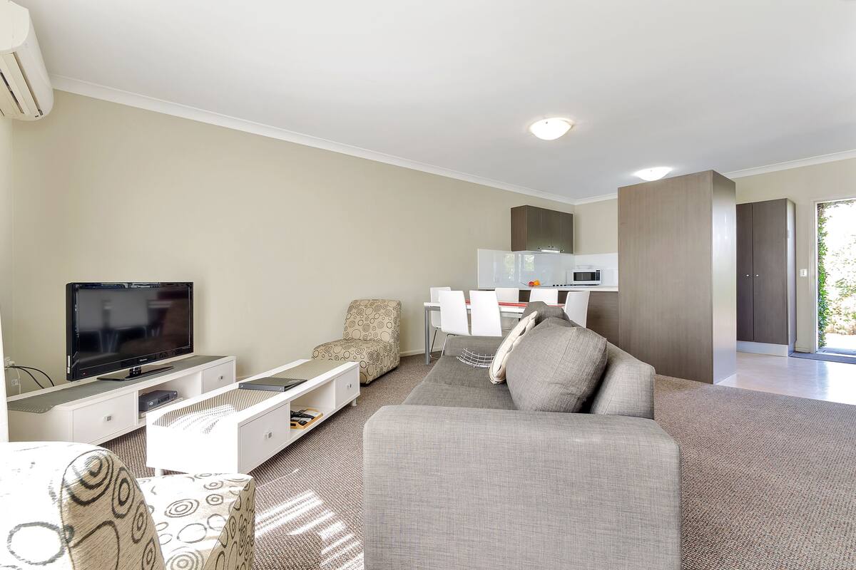 Una sala de estar moderna con televisor de pantalla plana, sofá gris, sillón estampado y una zona de comedor con sillas blancas y una mesa.
