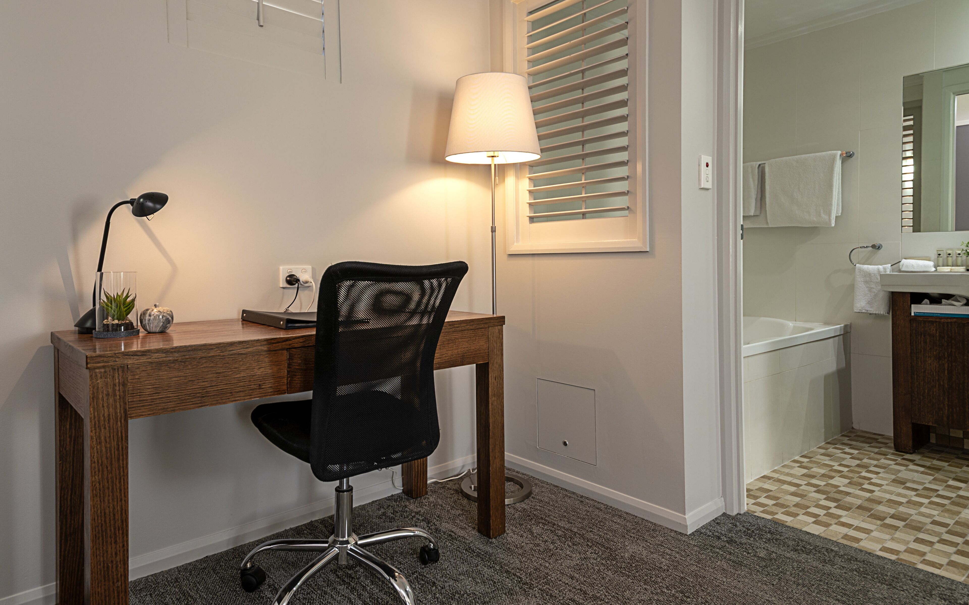Ein Arbeitsplatz mit Schreibtisch, Stuhl, Lampe und Blick ins Badezimmer.