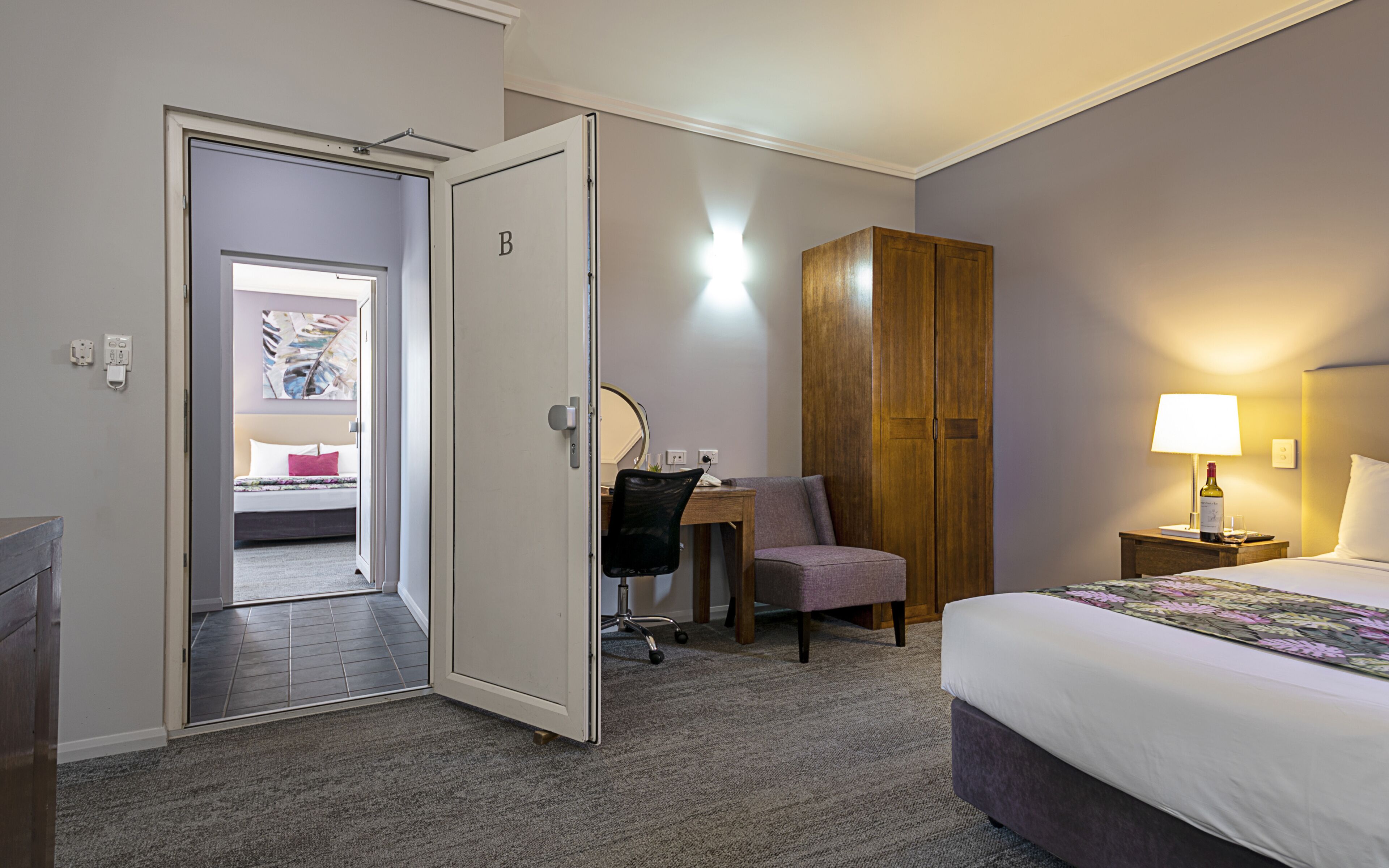 Une chambre d’hôtel avec un lit, un bureau, une chaise, une armoire et une salle de bain visible à travers une porte ouverte.