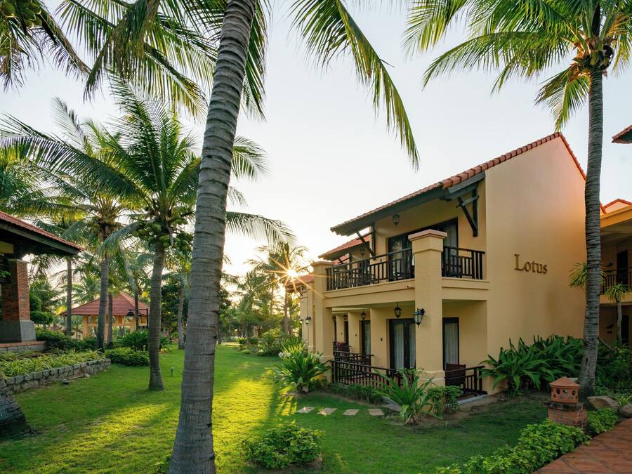 Un hotel de dos plantas, con balcón y un jardín bien cuidado con palmeras y arbustos.