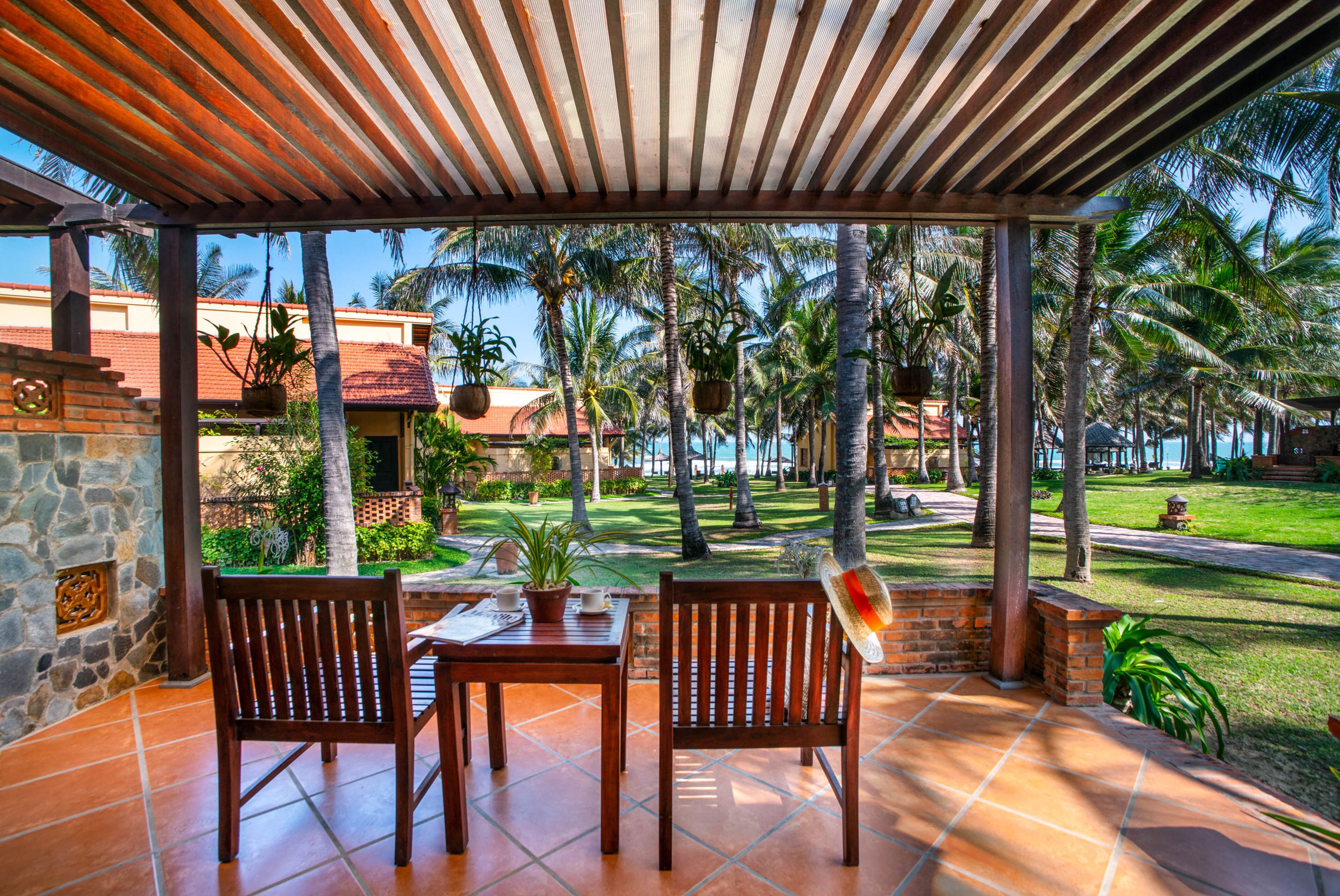 A covered outdoor seating area with two wooden chairs and a table, surrounded by palm trees and a view of a building with a red roof.