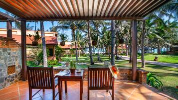 A covered outdoor seating area with two wooden chairs and a table, surrounded by palm trees and a view of a building with a red roof.