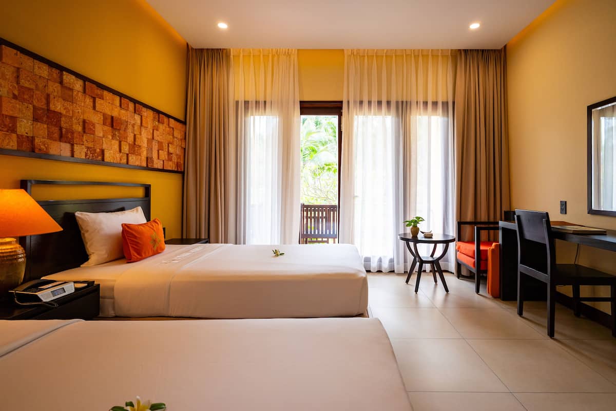 Une chambre d’hôtel avec deux lits, un bureau, une chaise, une petite table avec une plante et une vue sur la verdure à l’extérieur.