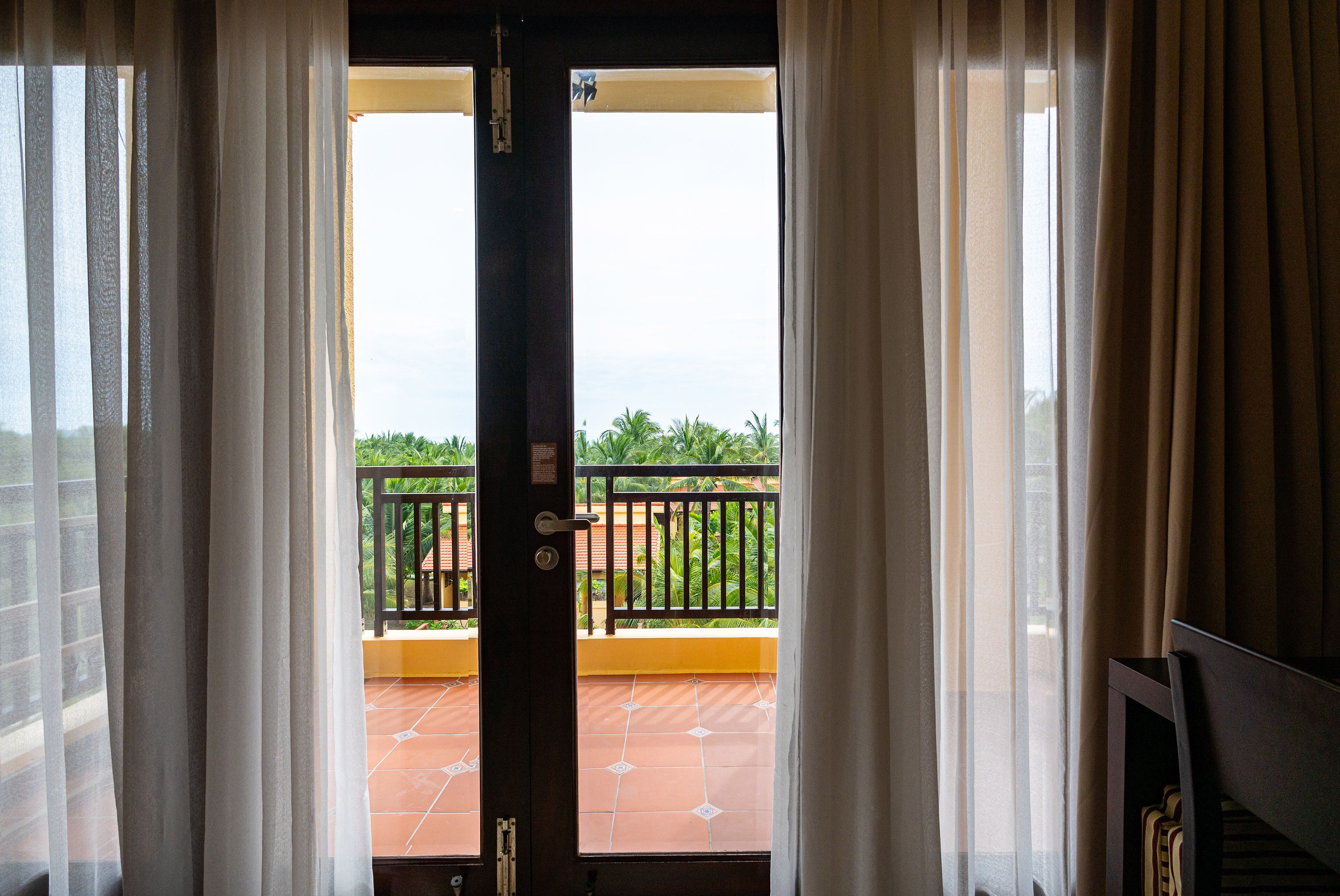 A view through a door with curtains, leading to a balcony with a tiled floor and a black railing.