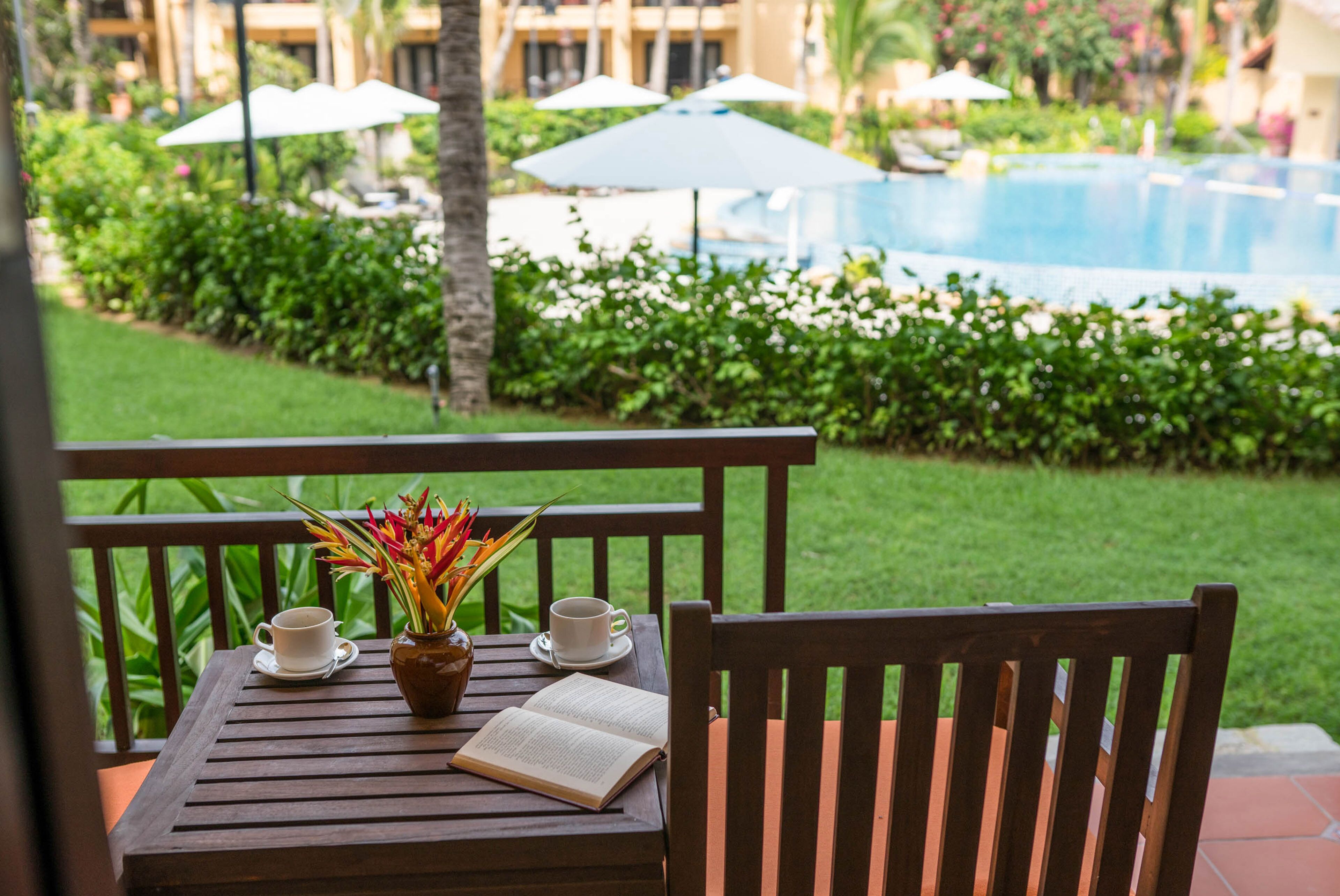 A wooden table with a book, two cups, and a vase with flowers on a balcony overlooking a pool.