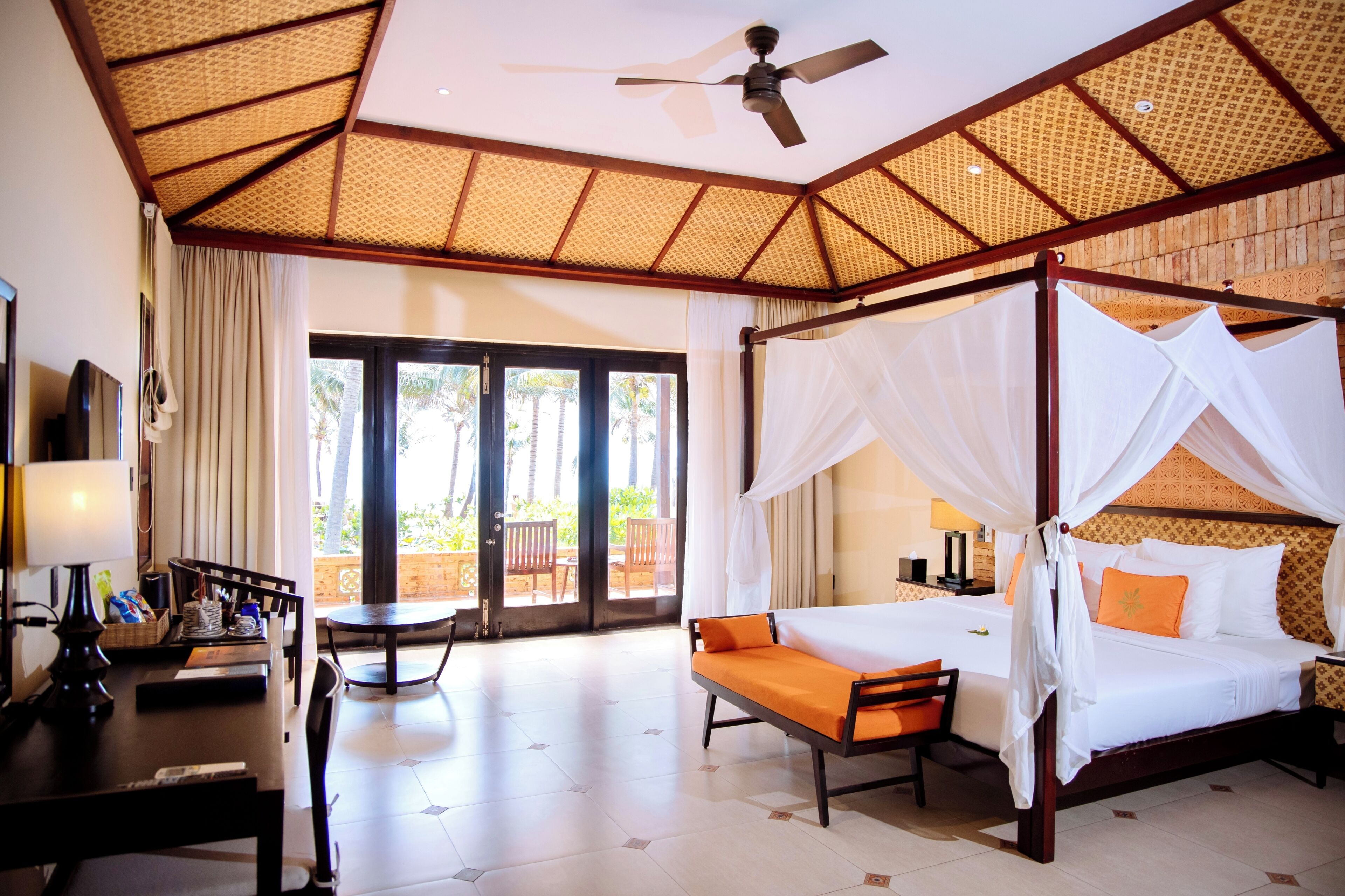 A room with a four-poster bed, a desk with a lamp, and a view through a glass door.