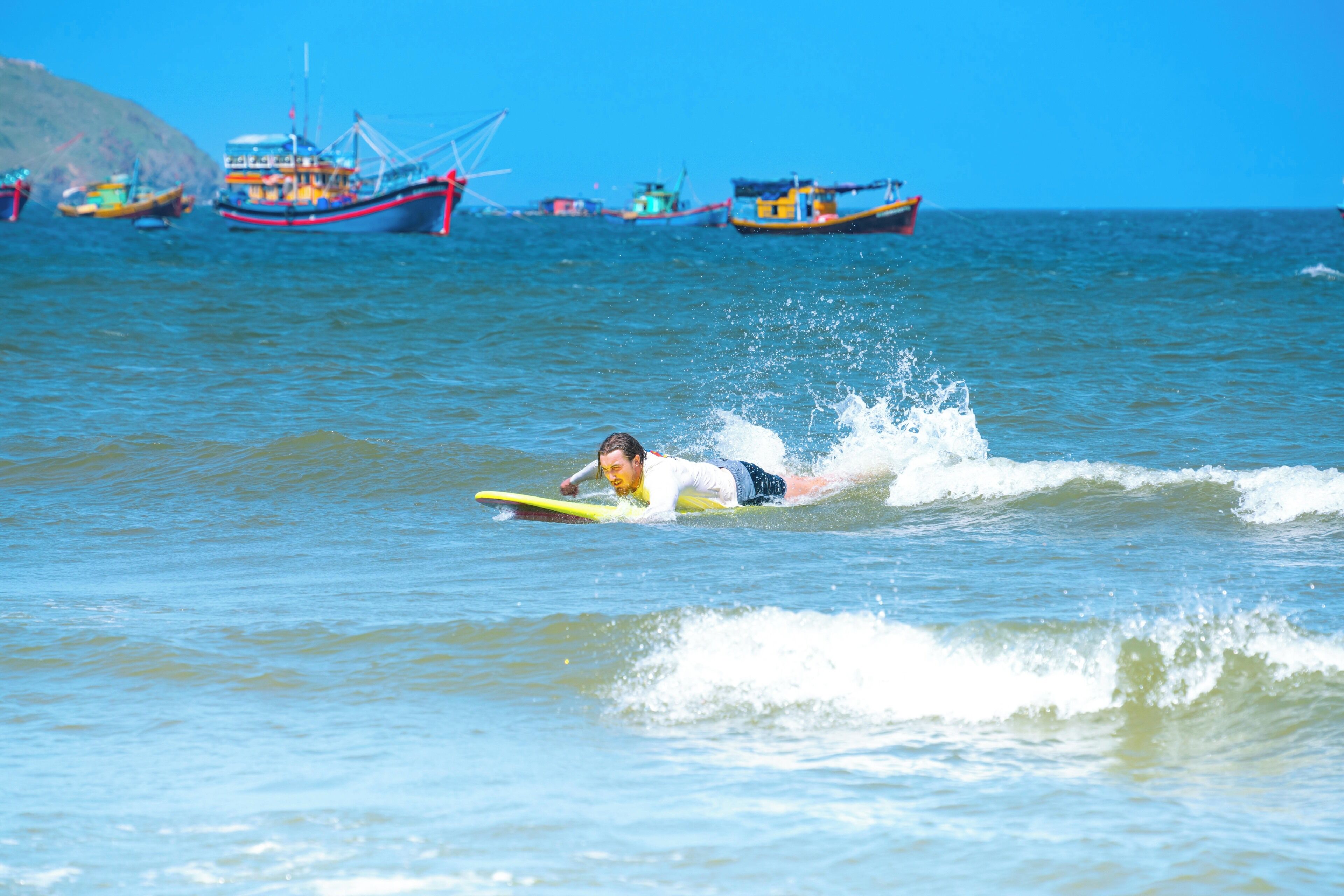A person surfing on a yellow surfboard in the ocean with several boats in the background.