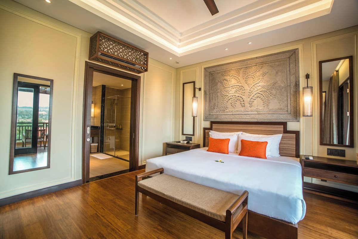 Une chambre d’hôtel avec un grand lit, un plancher en bois, un banc, un miroir, une table de chevet et un mur orné de motifs complexes.