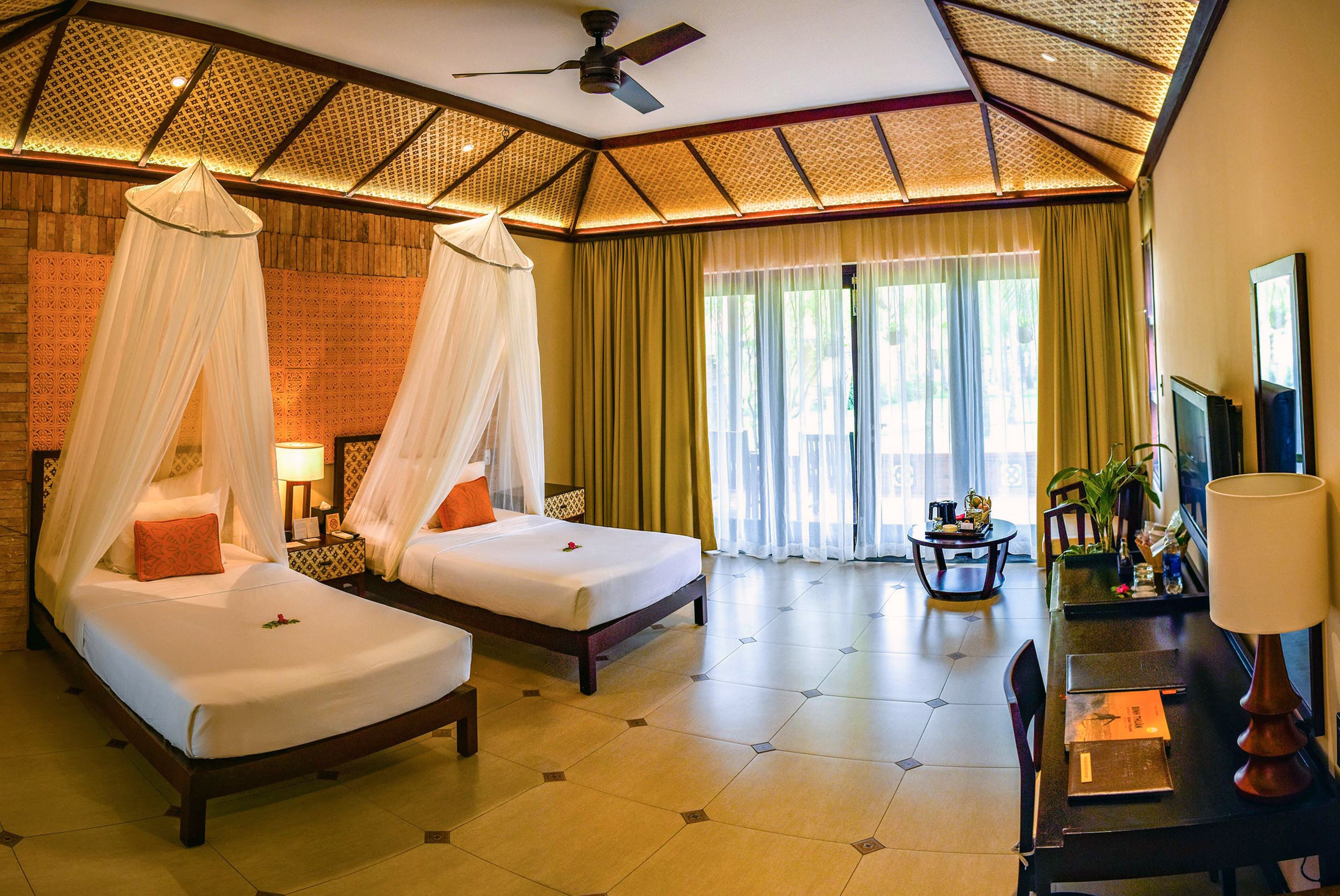 A hotel room with two beds, a desk, a chair, a TV, and a ceiling fan.