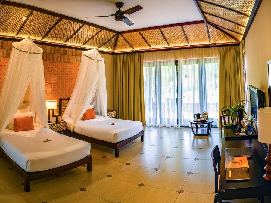 A hotel room with two beds, a desk, a chair, a TV, and a ceiling fan.