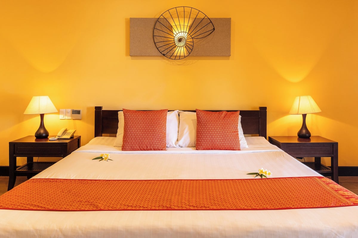 A hotel room with a large bed, two bedside tables with lamps, a wall-mounted decorative piece, and a flower on the bed.