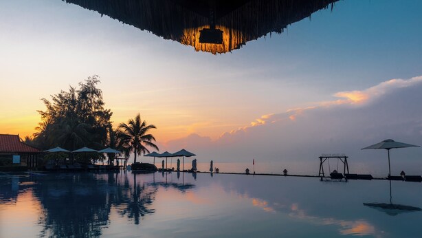 Un área de piscina tranquila con sombrillas y sillones reclinables, rodeada de palmeras y vistas al atardecer.
