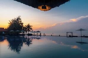 Un área de piscina tranquila con sombrillas y sillones reclinables, rodeada de palmeras y vistas al atardecer.