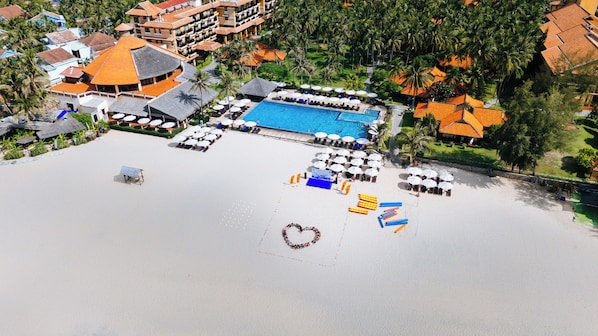 Vista aérea de un resort con piscina, tumbonas y un adorno con forma de corazón en la playa.
