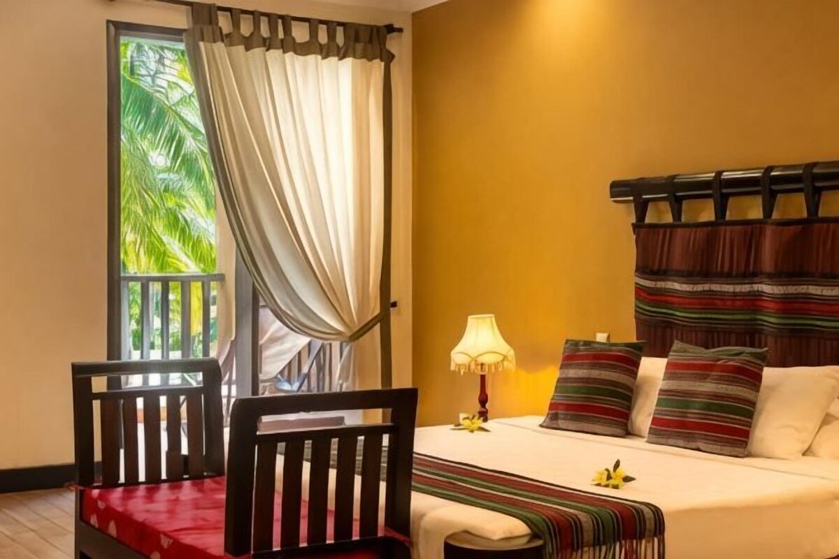 Un dormitorio con una cama, dos sillas, una lámpara y vistas a palmeras a través de una ventana.