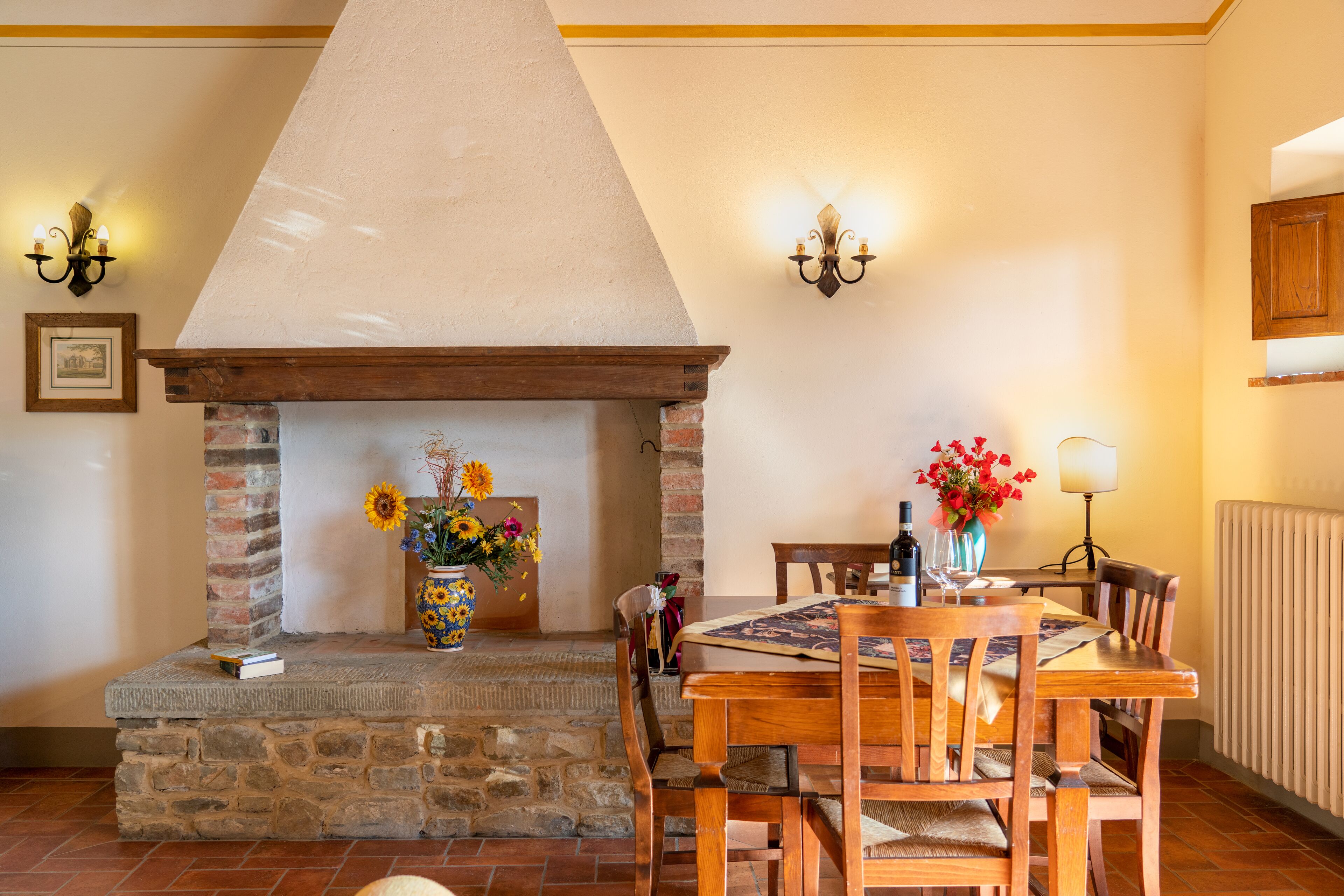 A dining area with a stone fireplace, wooden chairs, a table with a placemat, and a vase with flowers.