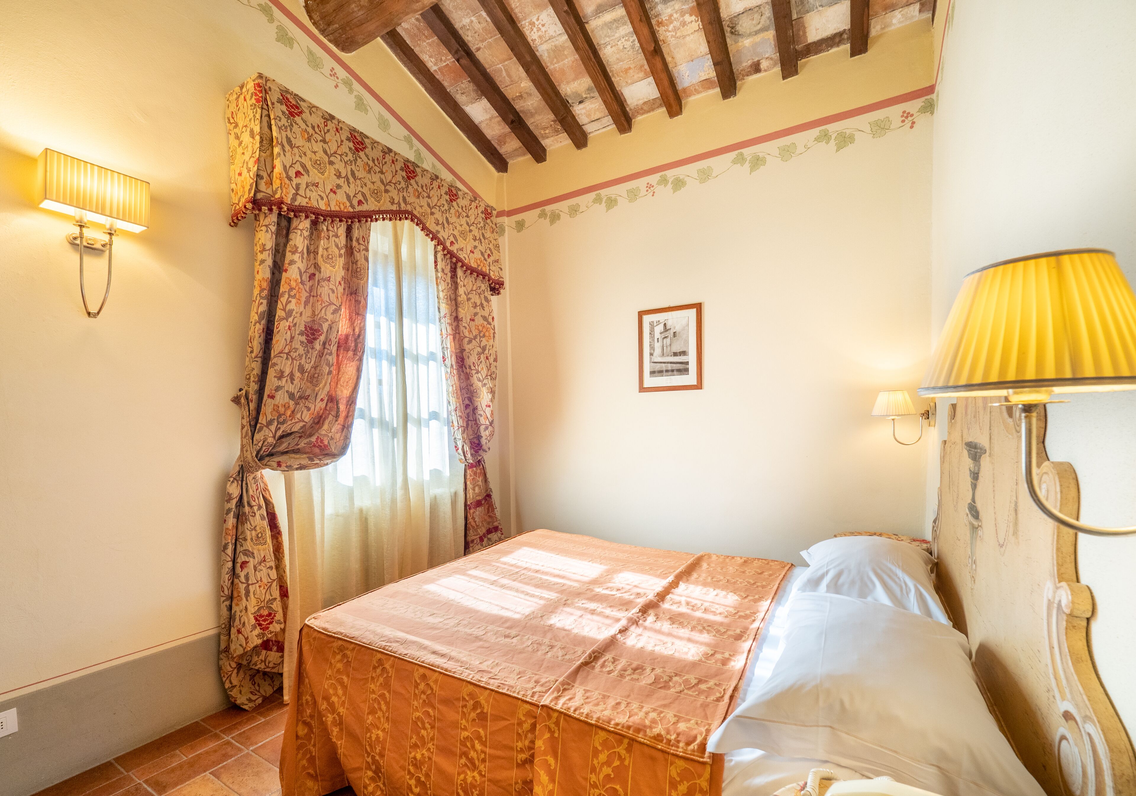 A bedroom with a bed, floral curtains, a wall-mounted lamp, and a framed picture.