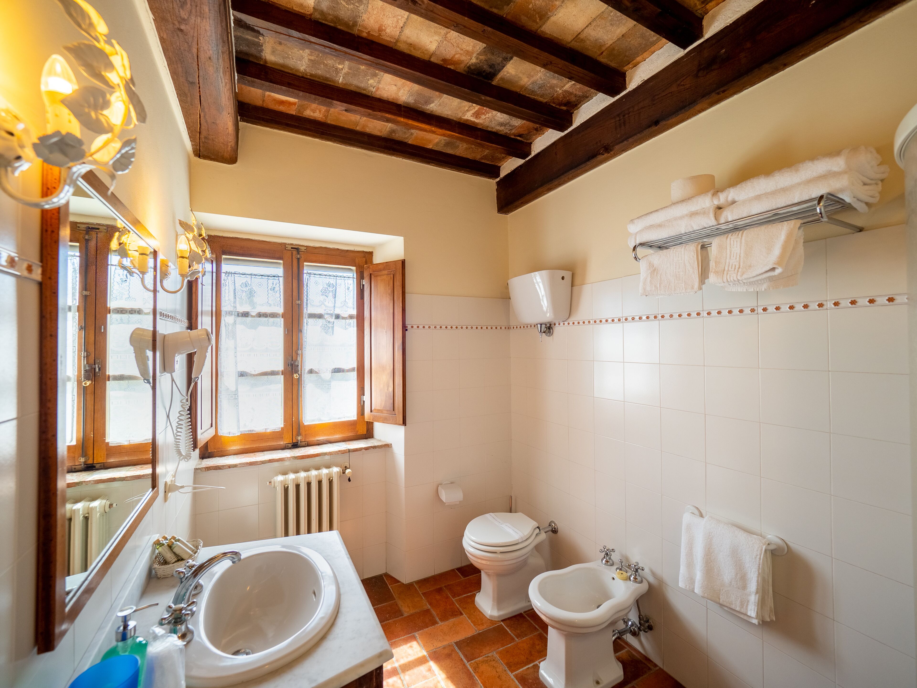 Et badeværelse med håndvask, to toiletter, et bidet, et håndklædestativ og et vindue med en radiator nedenunder.