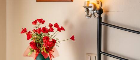 Una cama bien hecha con una manta estampada, un jarrón azul verdoso con flores rojas, un cuadro en la pared y una lámpara fijada a la pared.