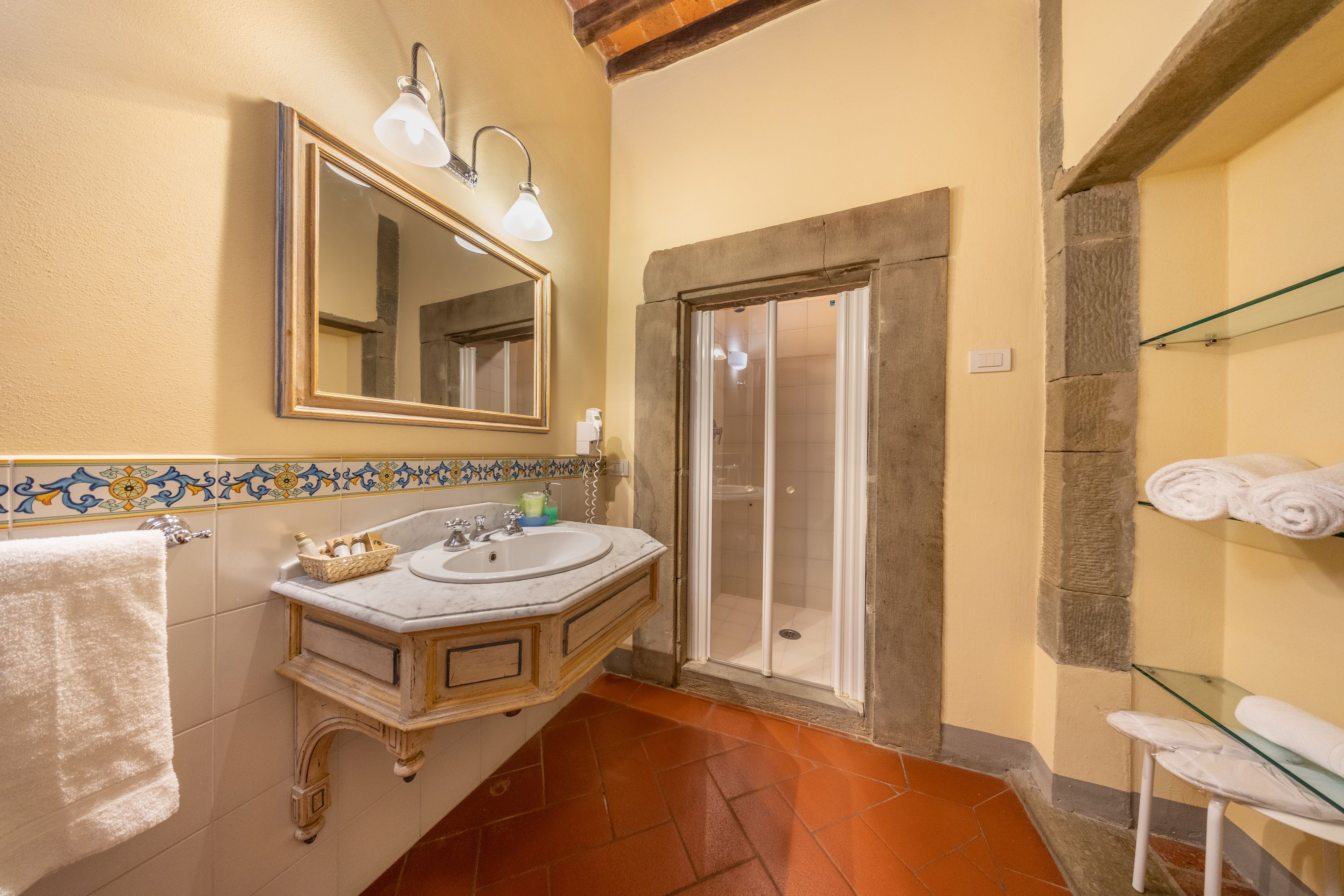 A bathroom with a marble sink, a mirror, a shower, and a glass shelf with towels.