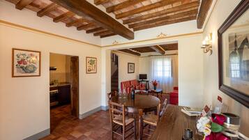 Uma área de refeições com uma mesa e cadeiras de madeira, uma cozinha ao fundo e uma escada que sobe.