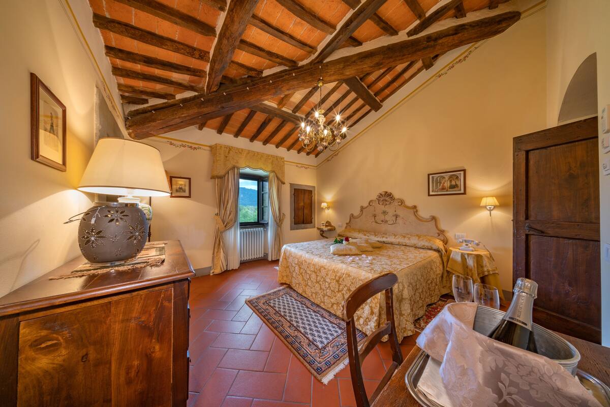 Une chambre avec un plafond en bois, un lit recouvert d’une couvre-lit à motifs floraux, une table de chevet avec une lampe et une fenêtre avec des rideaux.