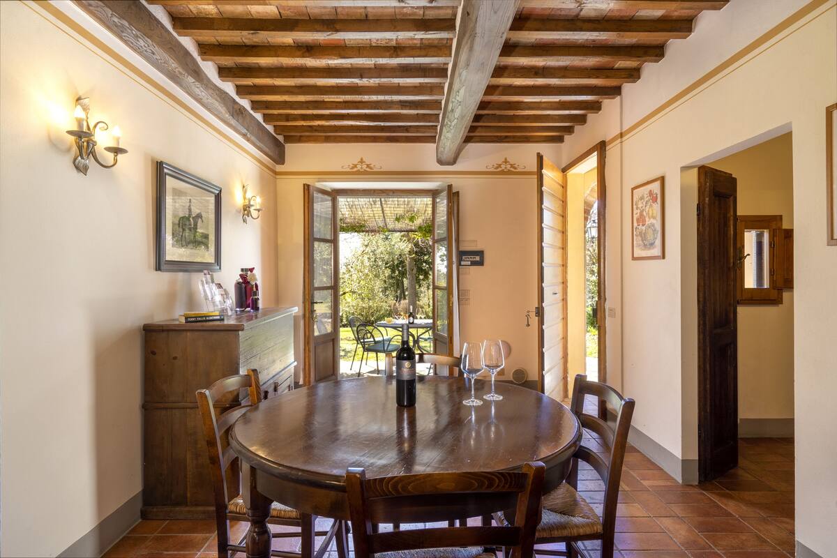 Un coin repas avec une table en bois, des chaises et une bouteille de vin. On y trouve également un buffet en bois et un tableau encadré accroché au mur.