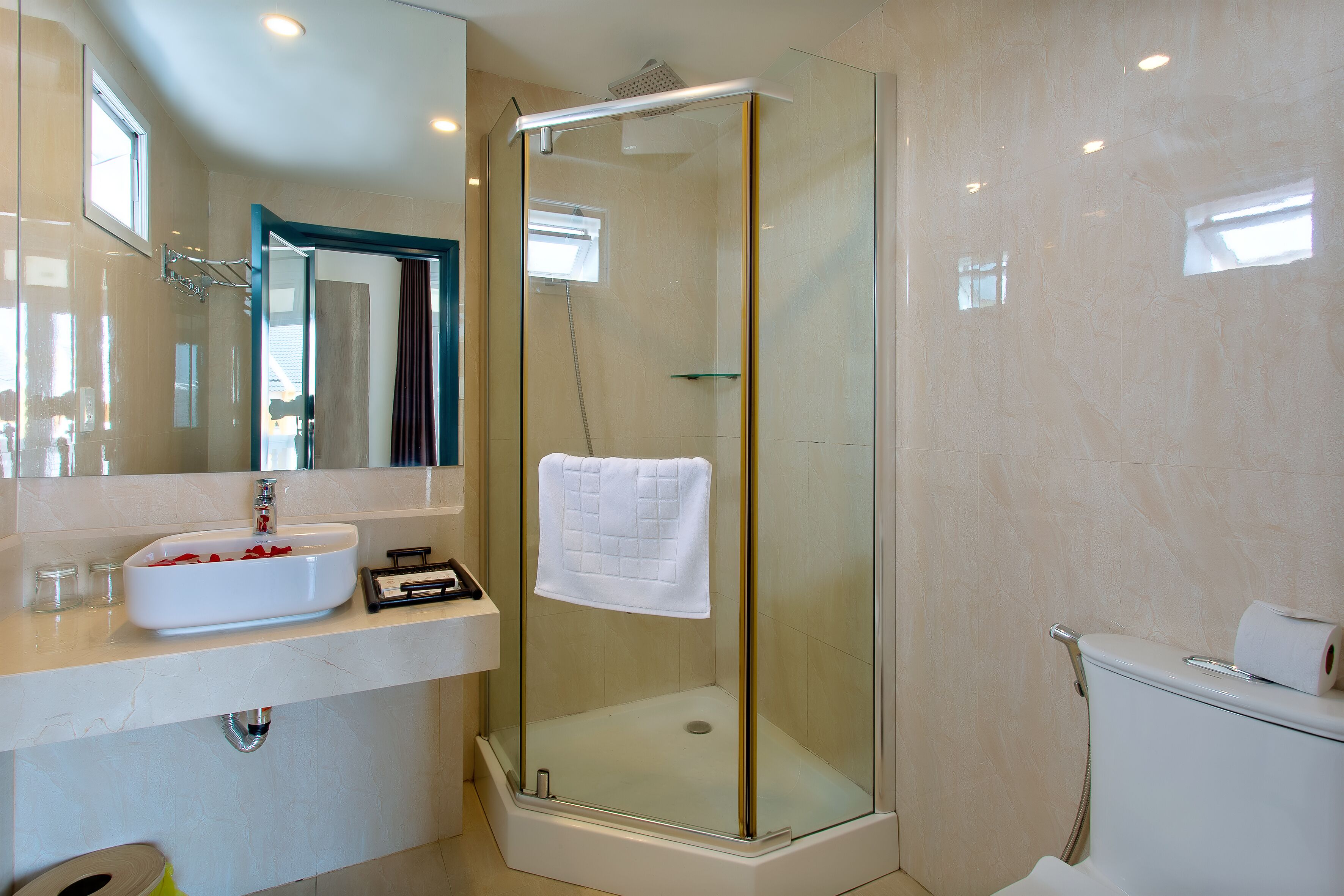 A modern bathroom with a glass-enclosed shower, a white sink with rose petals, a towel hanging on the shower door, and a toilet with a roll of toilet paper.
