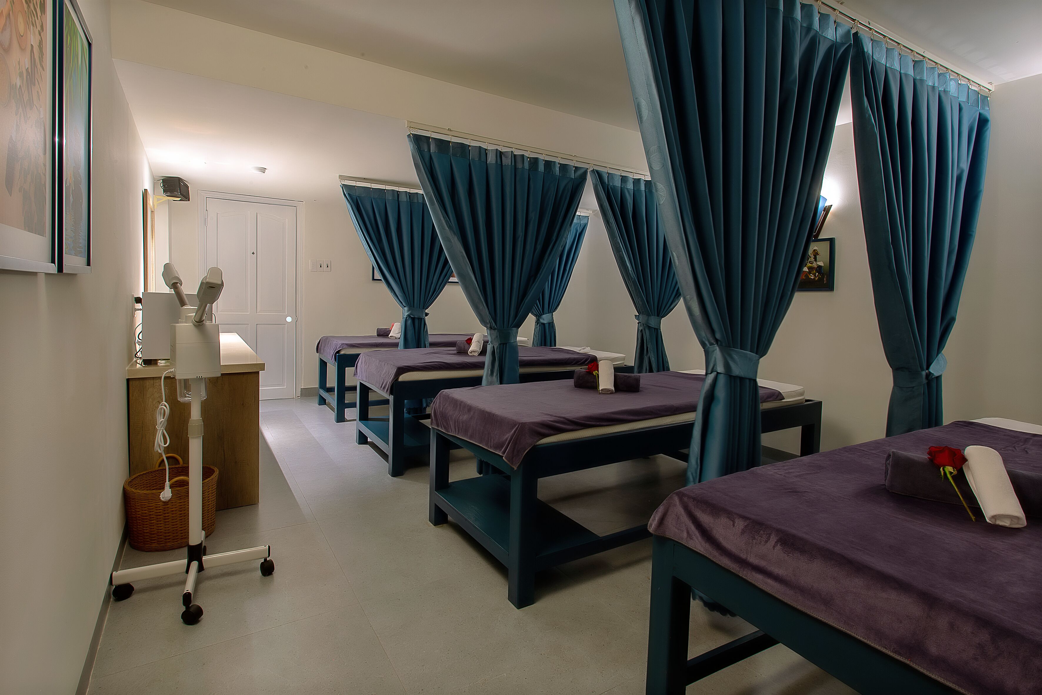 A room with four beds, each with a canopy and purple bedding. There is a small table with a lamp and a basket on the left.