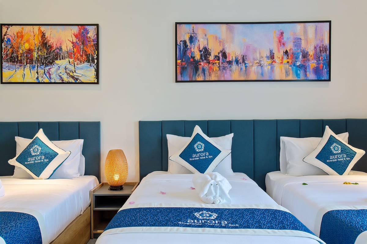 Quarto de hotel com duas camas, cada uma com almofada e manta estampada com o logotipo do hotel. Há um quadro na parede acima das camas, e uma mesa de cabeceira com abajur entre elas.