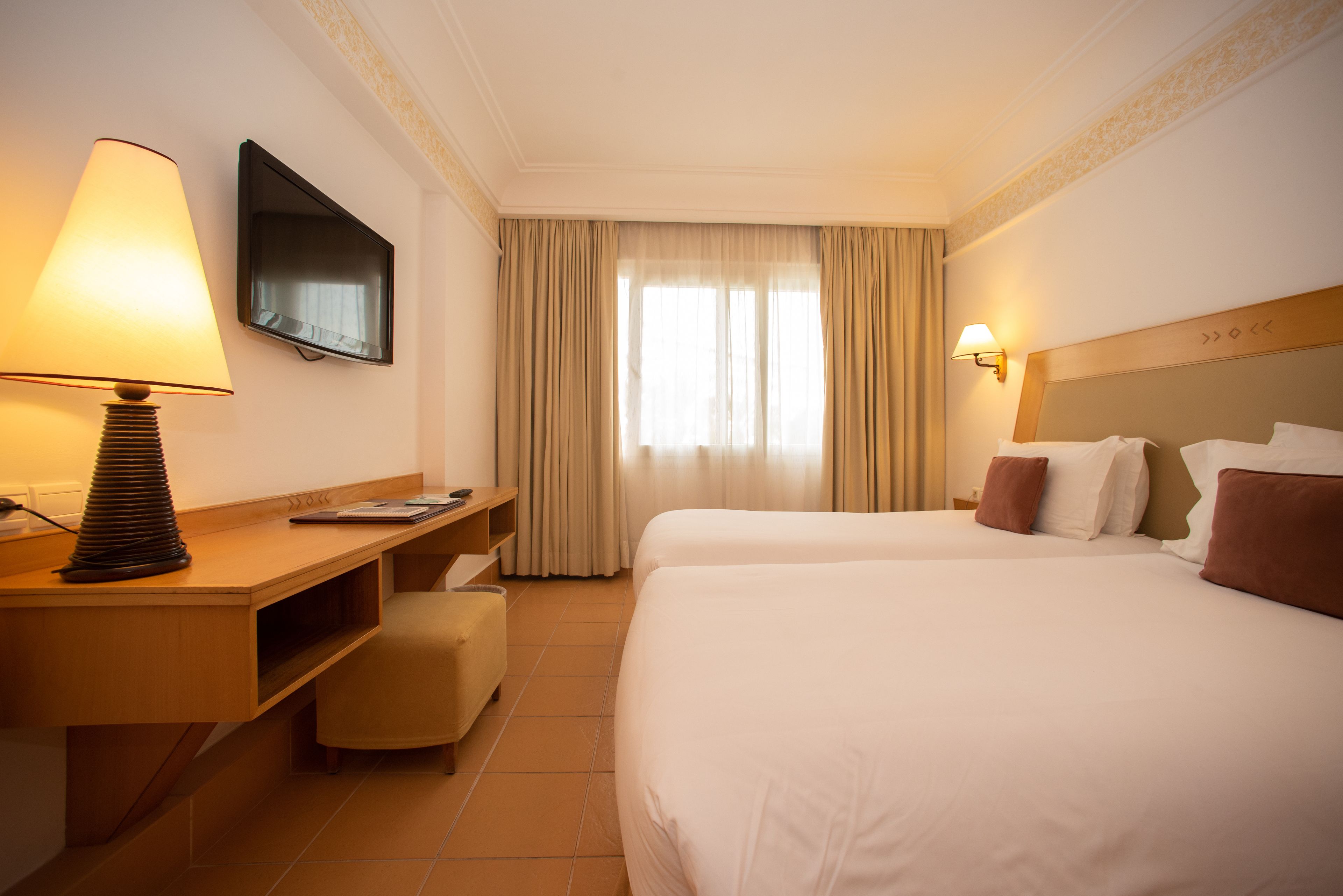 A hotel room with two beds, a desk, a chair, a TV, and a window with curtains.
