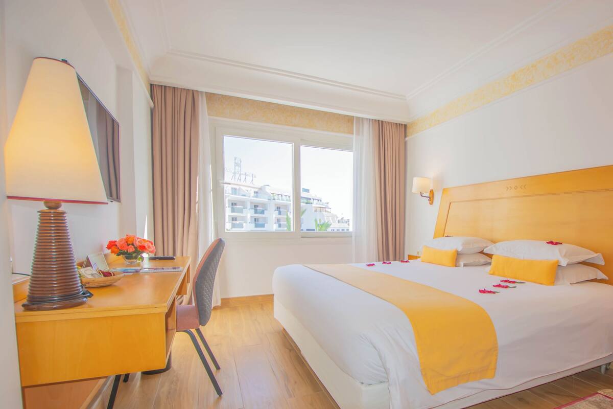 A hotel room with a large bed, a desk with a lamp, a chair, and a window with curtains.