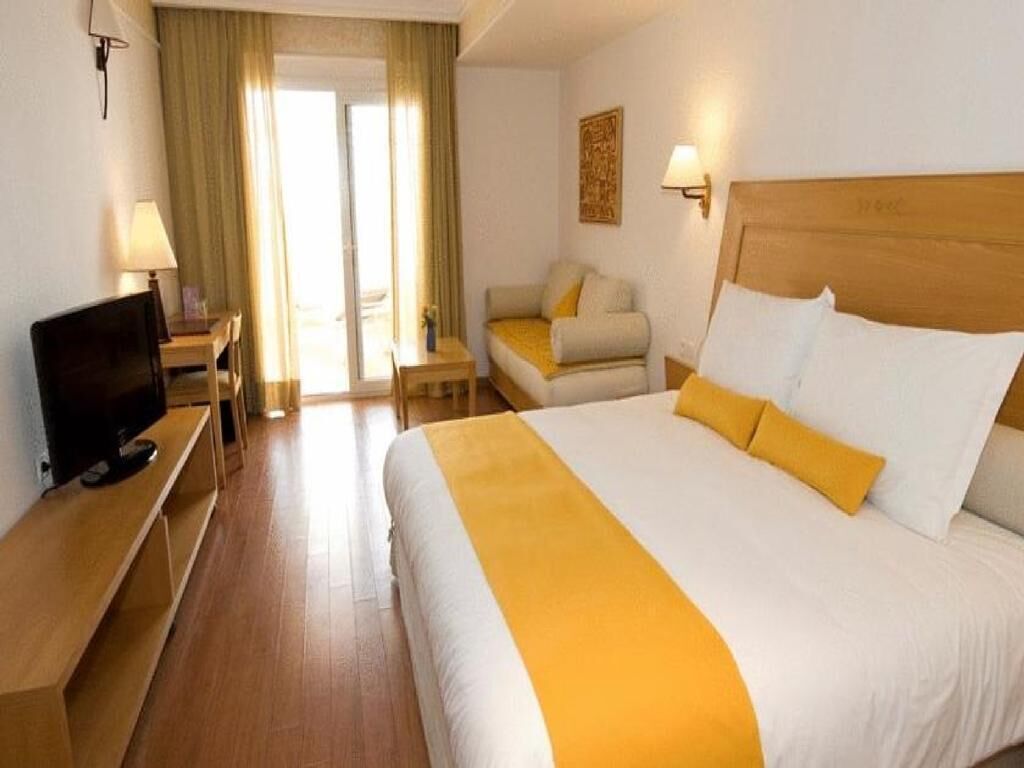 A hotel room with a bed, a television on a stand, a desk with a lamp, and a chair.