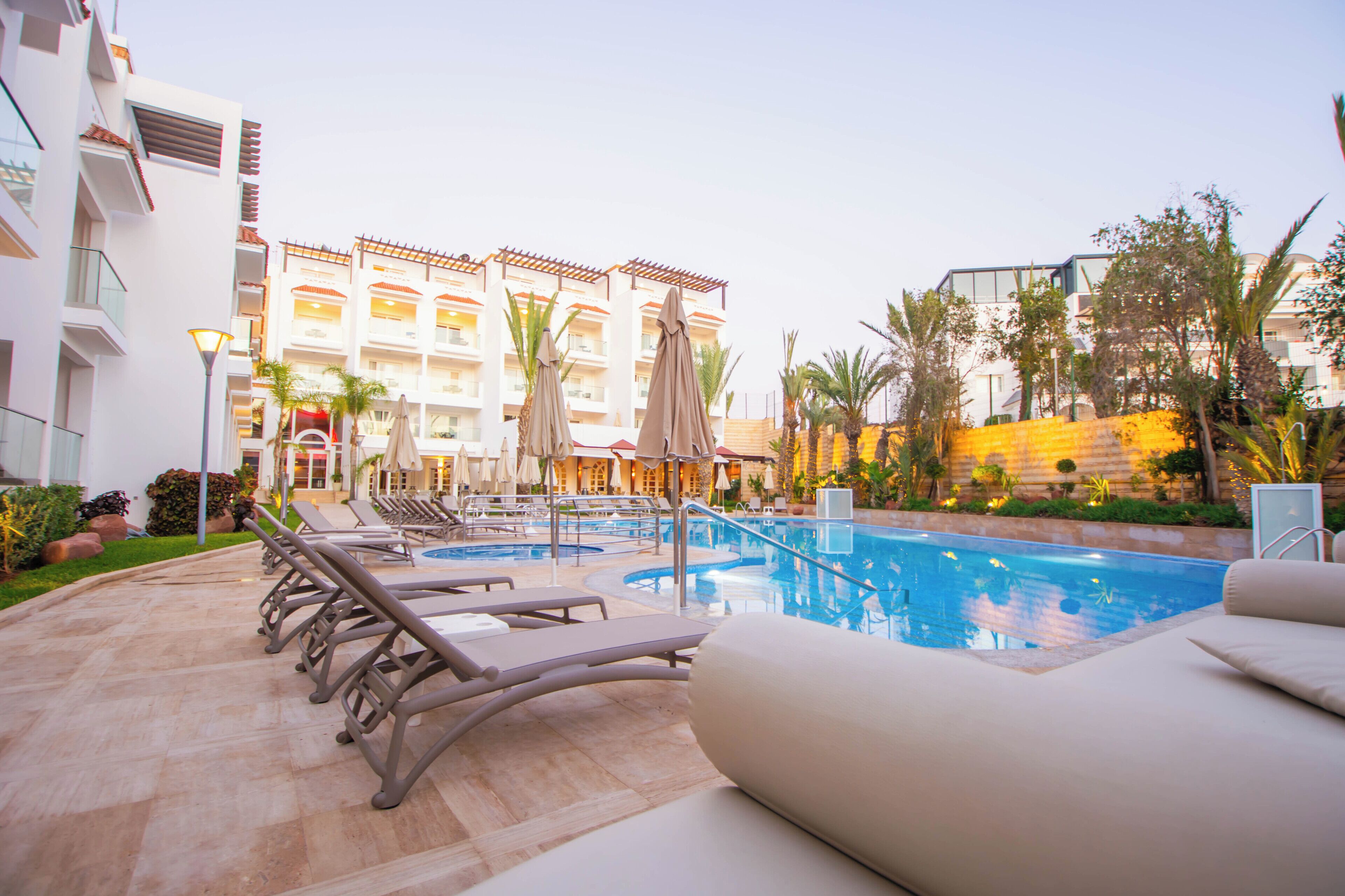 A hotel pool area with sun loungers and umbrellas, surrounded by palm trees and a building with multiple balconies.